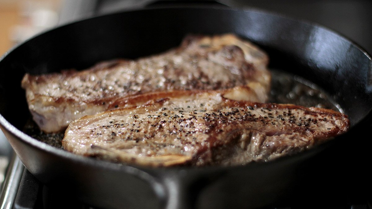 New York strip steaks cooking in a cast iron pan on a natural gas stove top