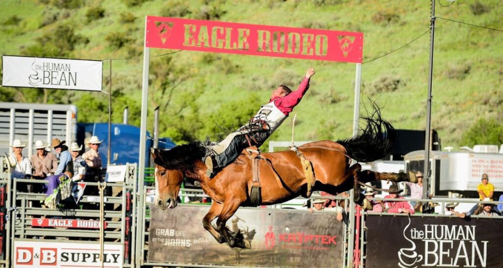 Cowboy riding a bucking bronc with The Eagle Rodeo banner in the background.