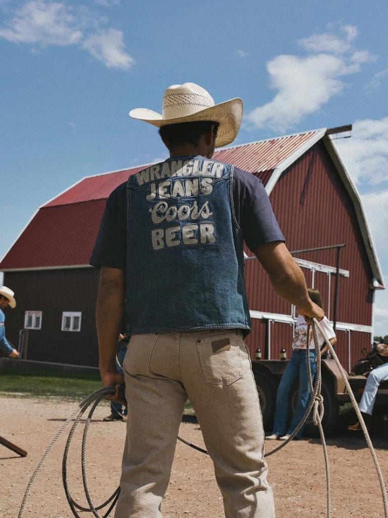 Man roping by a red barn while wearing a Wrangler Jean Coors Beer Vest