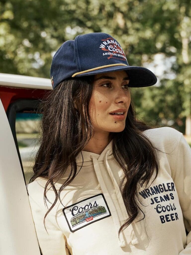 Woman wearing a denim blue Wrangler Coors Buckin Cap and cream color Wrangler Coors Hoodie