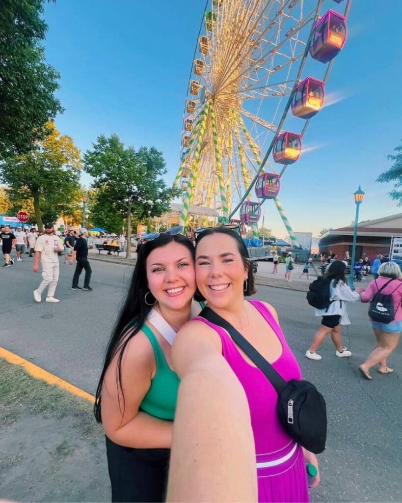 Two females enjoying themselves at Minnesota State Fair w Ferris wheel in the background.